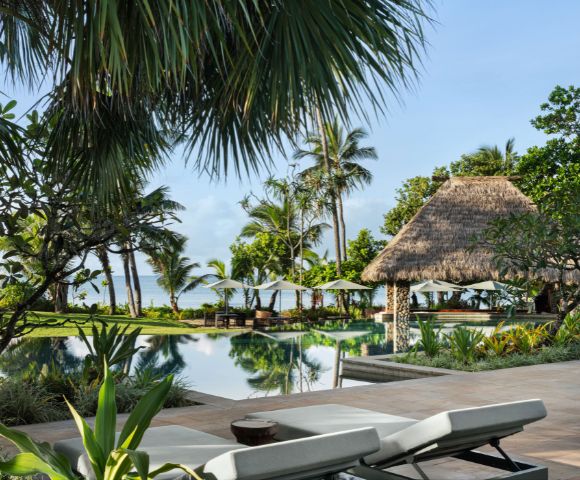 Tropical resort scene with palm trees, a tranquil pool, and a thatched gazebo. Two white sun loungers in the foreground suggest a relaxing vibe.
