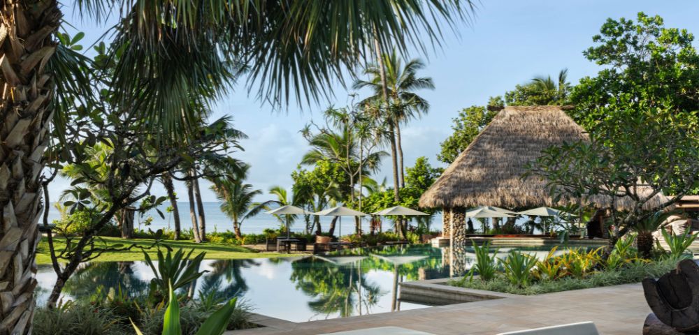 Tropical resort scene with palm trees, a tranquil pool, and a thatched gazebo. Two white sun loungers in the foreground suggest a relaxing vibe.