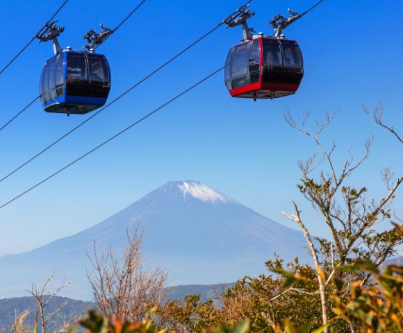 Two cable cars, one blue and one red, glide above foliage with Mount Fuji in the background under a clear blue sky, conveying tranquility and adventure.