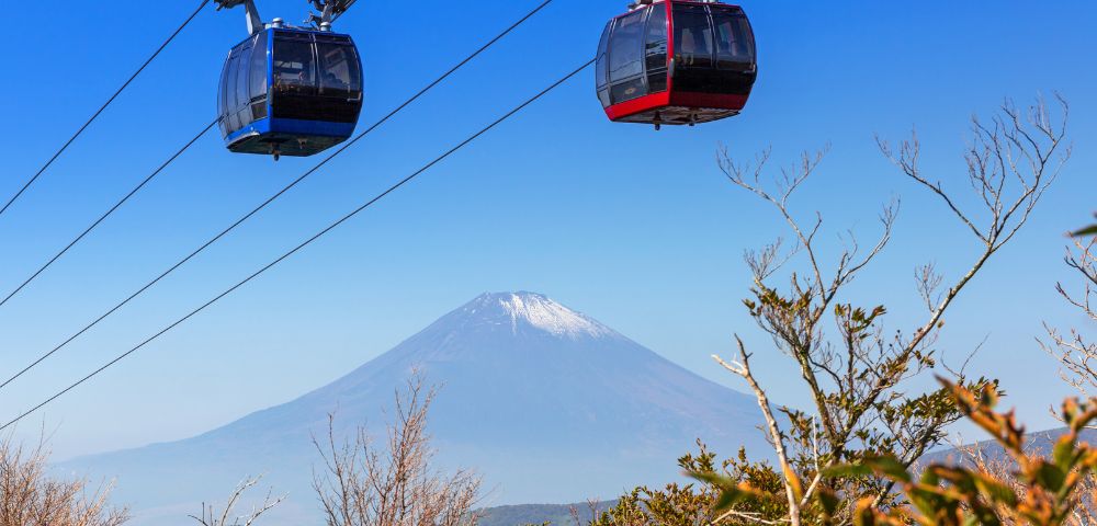 Two cable cars, one blue and one red, glide above foliage with Mount Fuji in the background under a clear blue sky, conveying tranquility and adventure.
