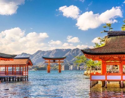 A serene scene at Itsukushima Shrine with red wooden buildings and a floating torii gate in calm water, set against mountains and a blue sky with clouds.
