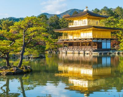 Golden pavilion reflecting on tranquil pond, surrounded by lush trees and distant mountains under a clear blue sky, creating a serene atmosphere.