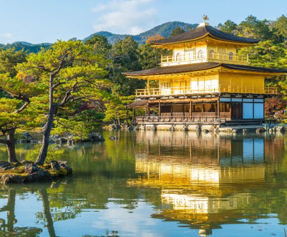 Golden pavilion reflecting on tranquil pond, surrounded by lush trees and distant mountains under a clear blue sky, creating a serene atmosphere.