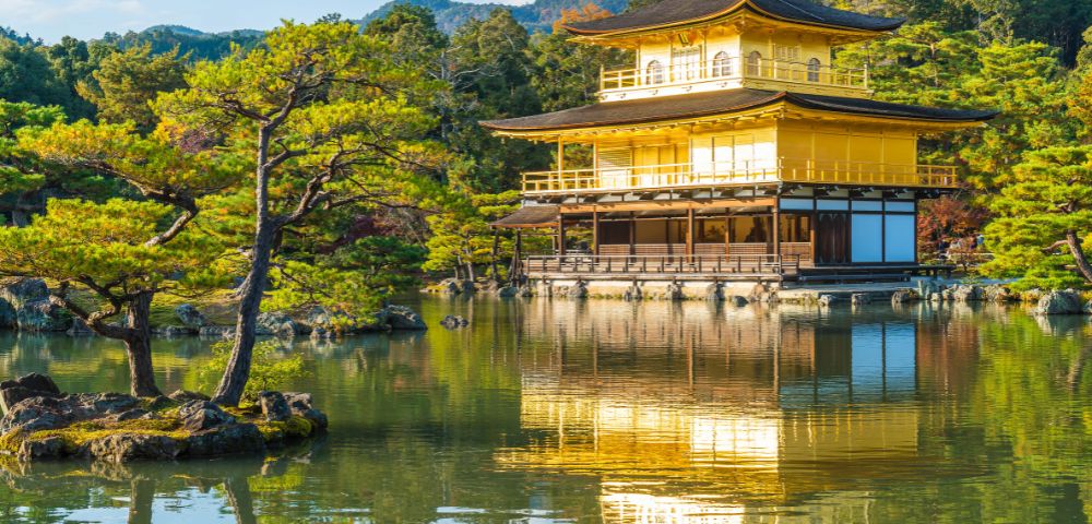 Golden pavilion reflecting on tranquil pond, surrounded by lush trees and distant mountains under a clear blue sky, creating a serene atmosphere.