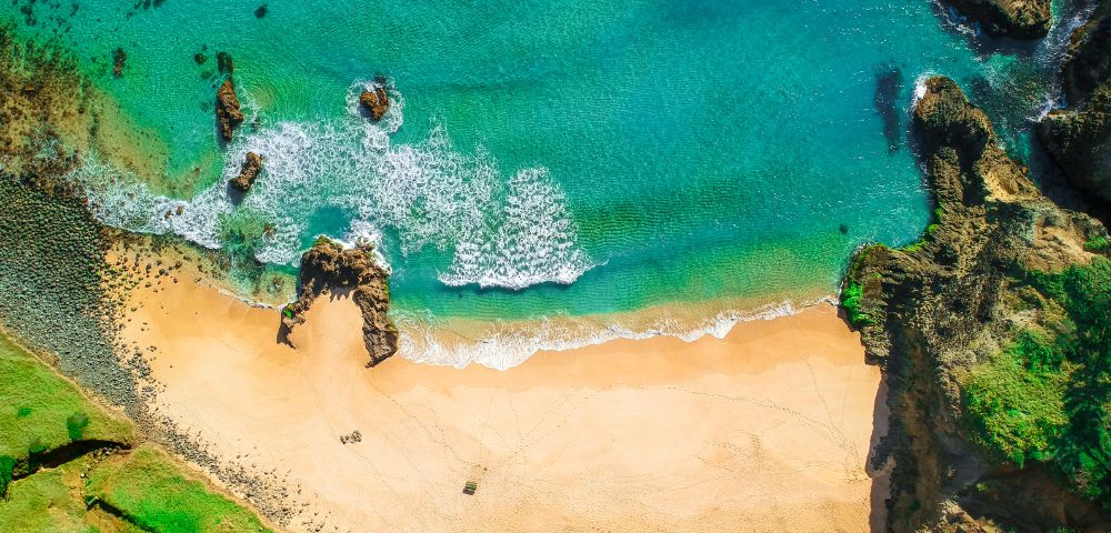 Aerial view of a serene beach with turquoise waves gently meeting golden sand, surrounded by rocky cliffs and lush greenery, creating a tranquil scene.