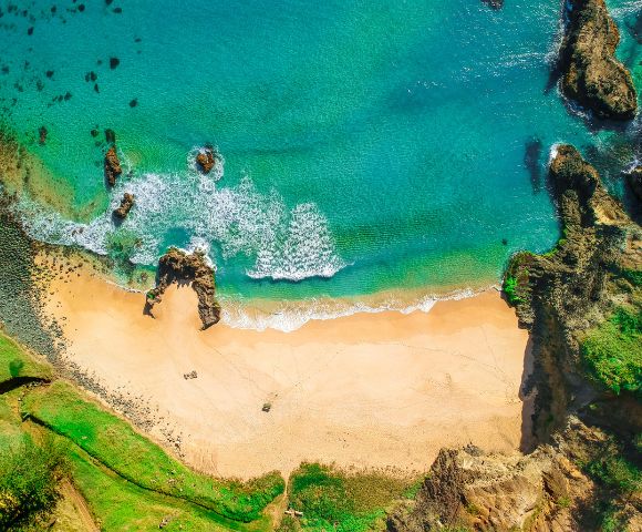 Aerial view of a serene beach with turquoise waves gently meeting golden sand, surrounded by rocky cliffs and lush greenery, creating a tranquil scene.
