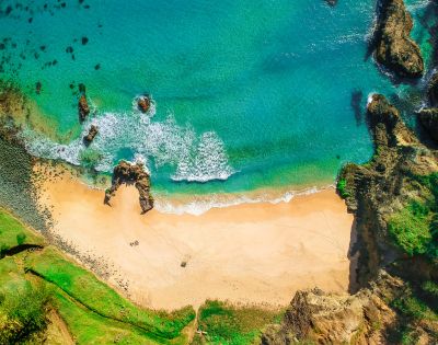 Aerial view of a serene beach with turquoise waves gently meeting golden sand, surrounded by rocky cliffs and lush greenery, creating a tranquil scene.