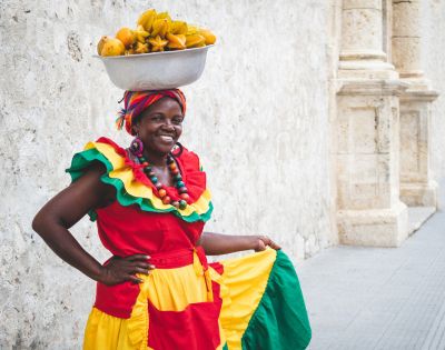 A smiling woman in vibrant, traditional clothing balances a bowl of fruit on her head. She stands against a textured stone wall, exuding joy and confidence.