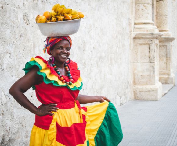 A smiling woman in vibrant, traditional clothing balances a bowl of fruit on her head. She stands against a textured stone wall, exuding joy and confidence.
