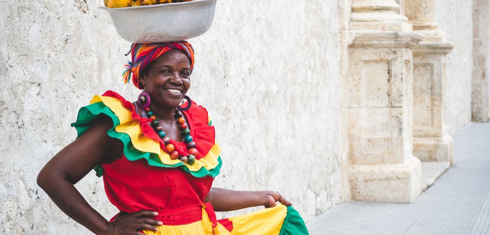 A smiling woman in vibrant, traditional clothing balances a bowl of fruit on her head. She stands against a textured stone wall, exuding joy and confidence.