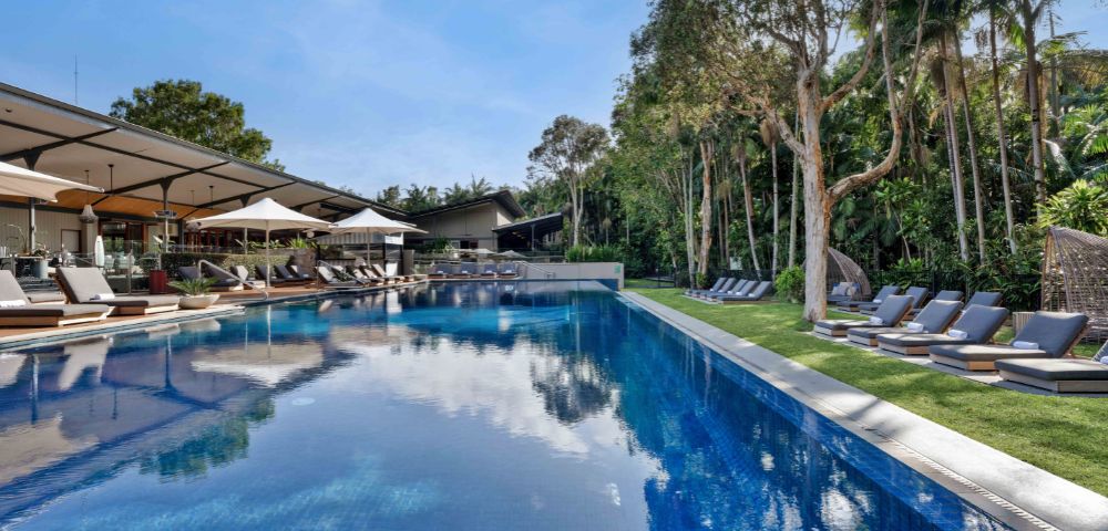 Luxurious poolside scene with clear blue water, surrounded by lounge chairs and lush trees. A serene atmosphere under a bright blue sky.