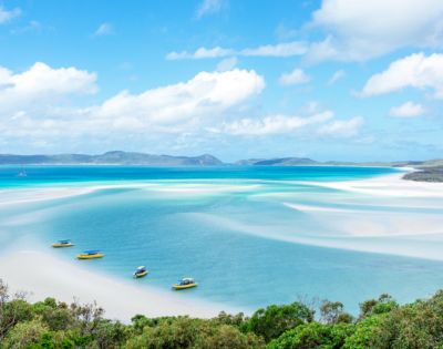 Aerial view of a serene beach with turquoise waters and white sandbanks. Four small boats are moored, with lush greenery in the foreground under a clear blue sky.