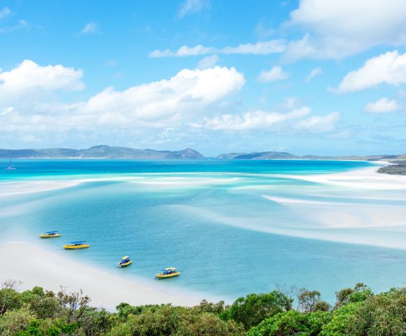 Aerial view of a serene beach with turquoise waters and white sandbanks. Four small boats are moored, with lush greenery in the foreground under a clear blue sky.