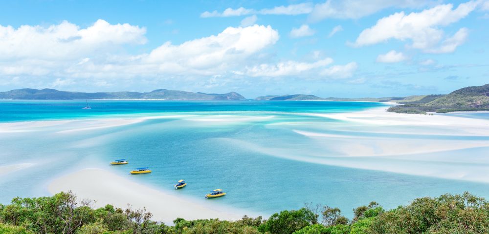 Aerial view of a serene beach with turquoise waters and white sandbanks. Four small boats are moored, with lush greenery in the foreground under a clear blue sky.
