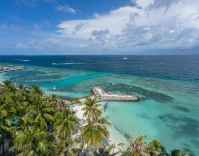 Aerial view of a tropical beach with turquoise waters and lush palm trees. A heart-shaped island and boats dot the sea, conveying a serene, tropical vibe.