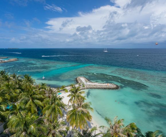 Aerial view of a tropical beach with turquoise waters and lush palm trees. A heart-shaped island and boats dot the sea, conveying a serene, tropical vibe.