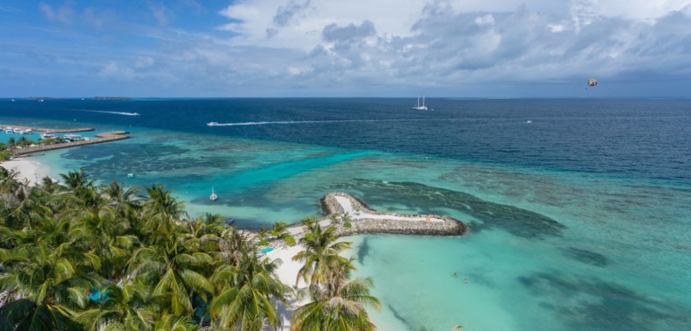 Aerial view of a tropical beach with turquoise waters and lush palm trees. A heart-shaped island and boats dot the sea, conveying a serene, tropical vibe.