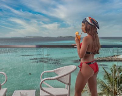 A woman in a red bikini stands on a balcony overlooking a tropical ocean view. She sips a drink, creating a relaxed and serene atmosphere.
