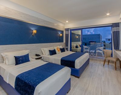 Modern hotel room with two double beds, blue and white decor, soft lighting, and a glass door leading to a balcony with chairs. Calm and inviting ambiance.