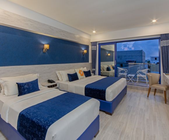 Modern hotel room with two double beds, blue and white decor, soft lighting, and a glass door leading to a balcony with chairs. Calm and inviting ambiance.