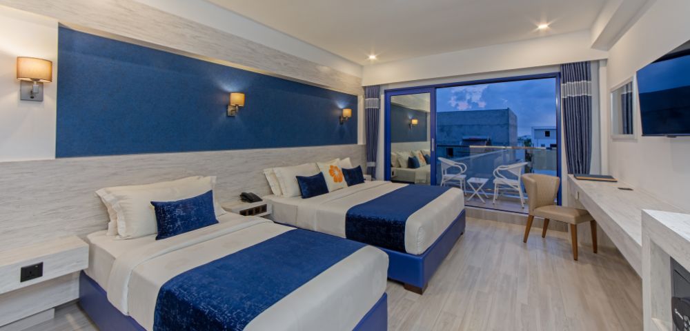 Modern hotel room with two double beds, blue and white decor, soft lighting, and a glass door leading to a balcony with chairs. Calm and inviting ambiance.