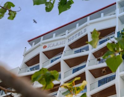 Modern white hotel building with angular balconies, seen from below against a blue sky. Green leaves frame the image, conveying a serene atmosphere.
