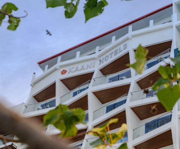 Modern white hotel building with angular balconies, seen from below against a blue sky. Green leaves frame the image, conveying a serene atmosphere.
