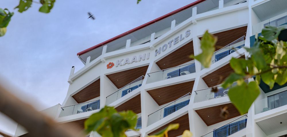 Modern white hotel building with angular balconies, seen from below against a blue sky. Green leaves frame the image, conveying a serene atmosphere.