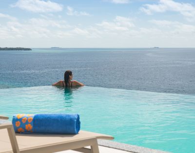 A person relaxes at the edge of an infinity pool overlooking a serene ocean under a partly cloudy sky. A lounger with a blue towel is in the foreground.
