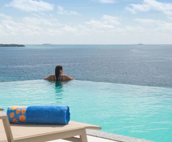 A person relaxes at the edge of an infinity pool overlooking a serene ocean under a partly cloudy sky. A lounger with a blue towel is in the foreground.