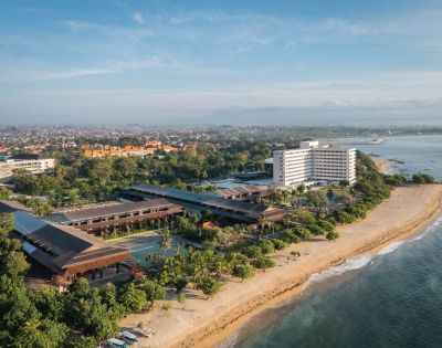 Aerial view of a beachfront hotel with modern architecture surrounded by lush greenery. The ocean is on the right, and a cityscape is visible in the distance.