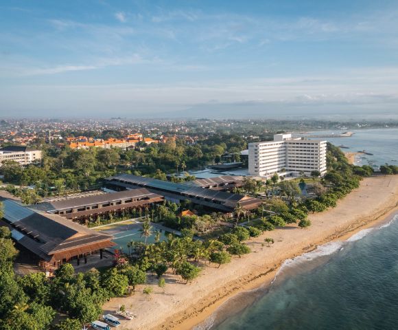 Aerial view of a beachfront hotel with modern architecture surrounded by lush greenery. The ocean is on the right, and a cityscape is visible in the distance.