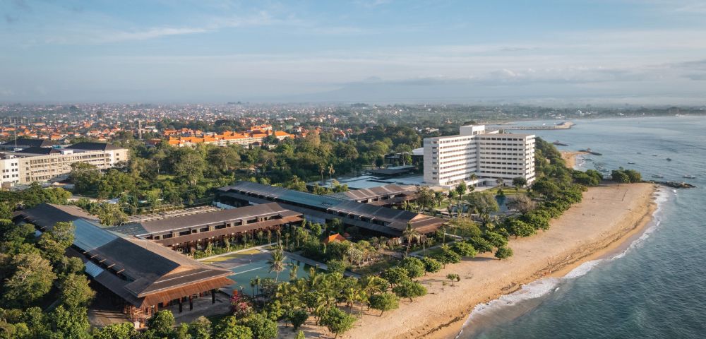 Aerial view of a beachfront hotel with modern architecture surrounded by lush greenery. The ocean is on the right, and a cityscape is visible in the distance.