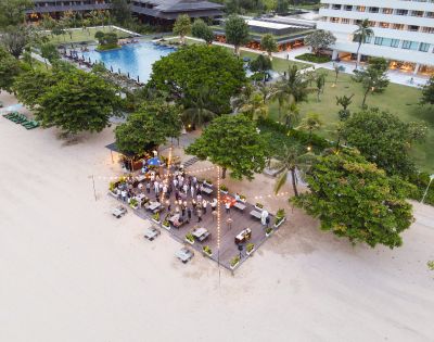 Aerial view of a beachside party with guests gathered on a deck under string lights. Nearby, a pool and hotel are surrounded by lush greenery. Festive and relaxed ambiance.