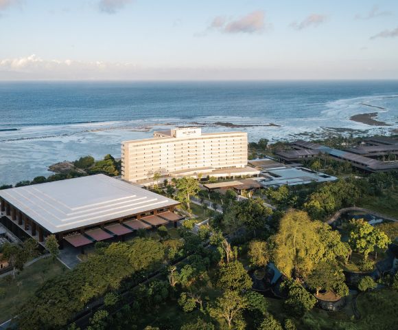 Aerial view of a seaside resort with a modern white hotel, surrounded by lush greenery and overlooking a calm ocean under a partly cloudy sky.