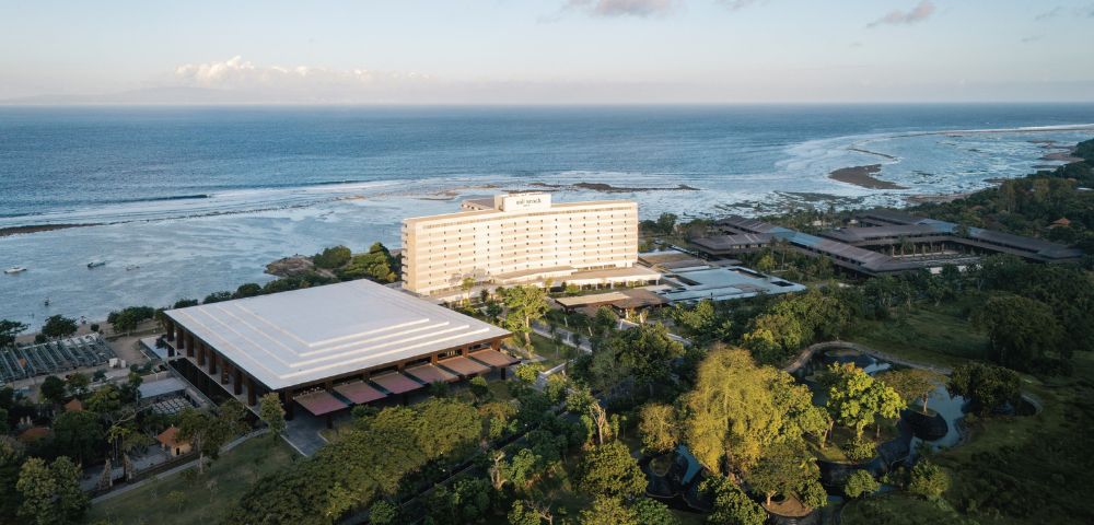 Aerial view of a seaside resort with a modern white hotel, surrounded by lush greenery and overlooking a calm ocean under a partly cloudy sky.