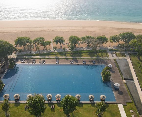 Aerial view of a large, serene pool surrounded by sun loungers under umbrellas. Beyond, a sandy beach and calm turquoise sea shimmer in sunlight.