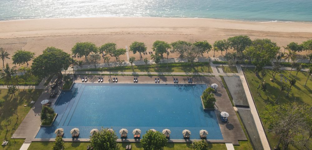 Aerial view of a large, serene pool surrounded by sun loungers under umbrellas. Beyond, a sandy beach and calm turquoise sea shimmer in sunlight.