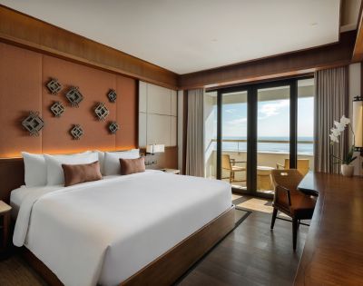 Elegant hotel room with a large bed, decorative pillows, and wall art. Glass doors open to a balcony with ocean views, creating a serene atmosphere.