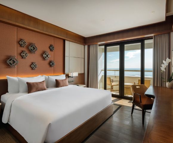 Elegant hotel room with a large bed, decorative pillows, and wall art. Glass doors open to a balcony with ocean views, creating a serene atmosphere.