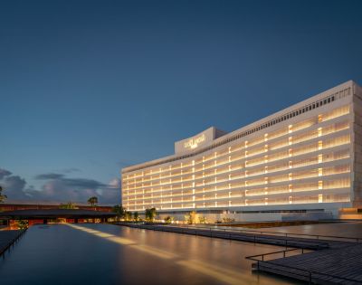 Elegant hotel building at dusk, with warm interior lights glowing. The structure reflects on a calm water feature, under a deep blue sky.