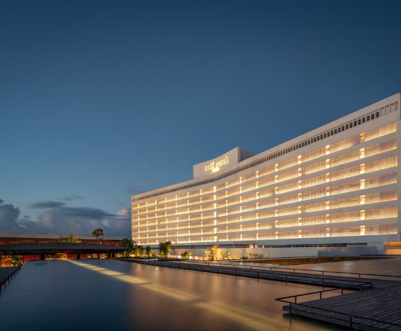 Elegant hotel building at dusk, with warm interior lights glowing. The structure reflects on a calm water feature, under a deep blue sky.