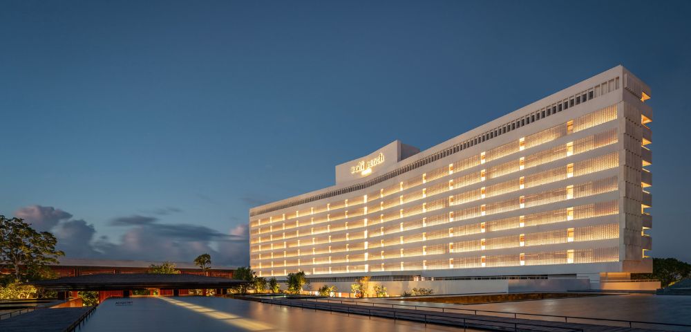 Elegant hotel building at dusk, with warm interior lights glowing. The structure reflects on a calm water feature, under a deep blue sky.