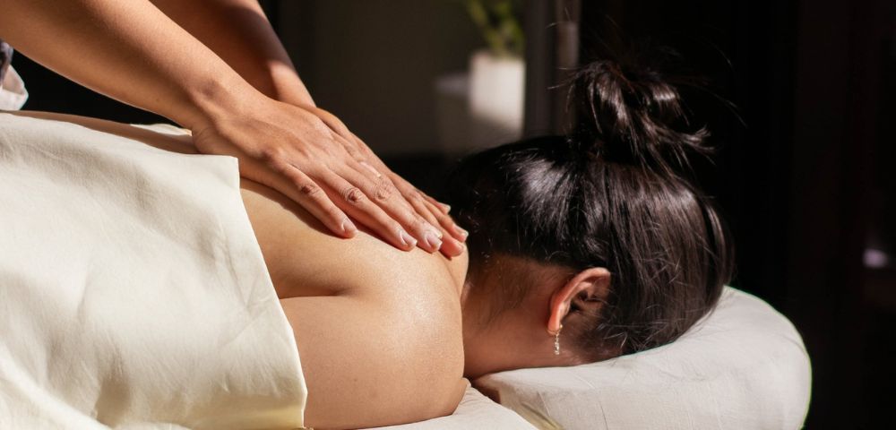 Woman receiving a relaxing back massage in dimly lit spa setting, conveying calm and serenity. A white towel covers her lower body.
