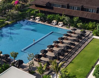Aerial view of a luxurious hotel pool with a single swimmer. Rows of elegant sun loungers and umbrellas line the deck. Lush greenery surrounds the area.