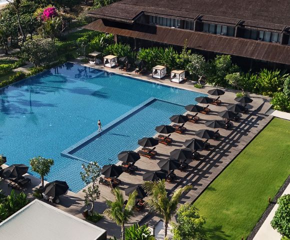 Aerial view of a luxurious hotel pool with a single swimmer. Rows of elegant sun loungers and umbrellas line the deck. Lush greenery surrounds the area.