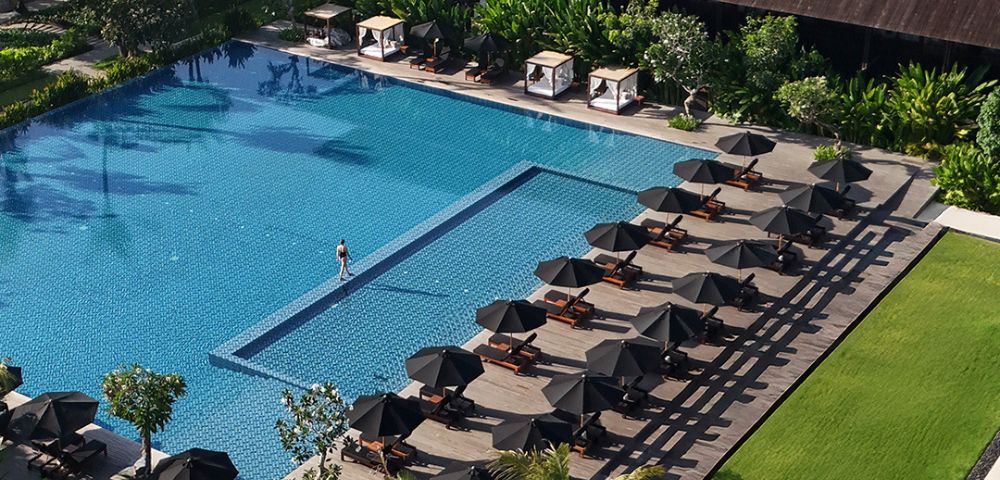 Aerial view of a luxurious hotel pool with a single swimmer. Rows of elegant sun loungers and umbrellas line the deck. Lush greenery surrounds the area.
