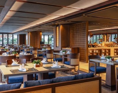 Spacious restaurant with elegant wooden decor, neatly set tables with white napkins, blue cushioned chairs, and large windows providing natural light.