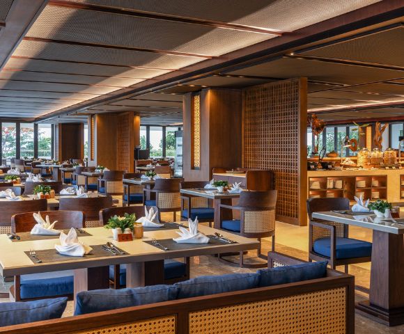 Spacious restaurant with elegant wooden decor, neatly set tables with white napkins, blue cushioned chairs, and large windows providing natural light.
