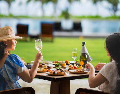 Two women seated at a table outdoors, enjoying a meal with a variety of dishes and wine. They appear relaxed, with a scenic pool and greenery in the background.